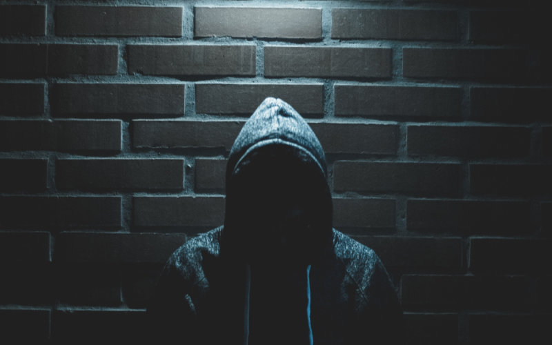 A hooded figure with their face hidden stands below a dim light