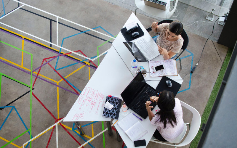 An aerial view of two women sitting at a desk with laptops and notepads