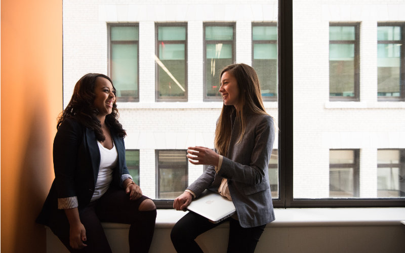 Two women sit on a window seat in an office window whilst talking to each other